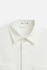 Sobrecamisa color beige de tejido elástico, con cuello solapa, manga corta y bolsillos de parche en el pecho. Cierre frontal con botones a presión.