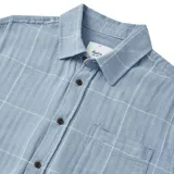 Camisa celeste con diseño a cuadros blancos, de manga corta, con cuello clásico, cierre frontal con botones marrones y bolsillo de parche en el pecho.