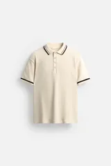Polo de hombre color crema, de corte regular, confeccionada en tejido de punto canalé. Presenta cuello solapa con cierre frontal de botones a presión y mangas cortas con ribete a contraste en color negro.