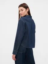 Camisa corta de jean azul oscuro, con cuello clásico, cierre frontal con botones, manga larga y bolsillo de parche en el pecho.