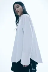 Camisa blanca oversized de popelina de algodón, con cuello clásico, botones frontales, hombros caídos, mangas largas con puños abotonados, canesú con pliegue en la espalda y bajo redondeado.