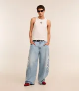Pantalón de jeans celeste de corte amplio, con cintura con trabillas, cierre tradicional y bolsillos. Presenta estampa lateral estilo maorí en color blanco.