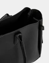 Cartera shopper de cuerina color negro con manijas superiores y charm a contratono. Tiene broche con botón para cerrar y cinturones laterales.