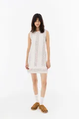 Vestido corto blanco con cuello redondo y sin mangas, confeccionado en tela ligera con detalles de encaje y puntilla.