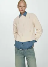 Sweater color crema de tejido de punto grueso con cuello redondo y manga larga. Presenta terminaciones de canalé y bordado de perlas en la parte delantera.
