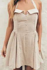Vestido corto a cuadros beige y blanco, con cuello halter que se ata en la nuca, escote recto con abertura y falda acampanada. Tiene bolsillos laterales ocultos, espalda elastizada y botones decorativos en el frente.