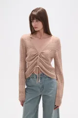 Sweater de poliéster color beige, con cuello en V, manga larga y frunces ajustables en el frente. Presenta un diseño de tejido calado.