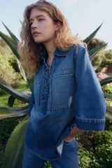 Camisa popover de denim azul con costuras en color contrastante, corte oversized, hombros caídos, cuello abierto con detalle de cordón, bolsillos grandes superiores, mangas al codo y bajo levemente redondeado.