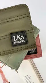 Billetera corta con cierre alrededor y tapa, confeccionada en tela capitoneada. Incluye varias reparticiones con tarjeteros y bolsillos internos, y una correa de mano desmontable. Disponible en negro, rojo y dorado.