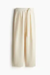 Pantalón sastrero color crudo, de tiro alto con elástico oculto en la cintura y cierre decorativo. Cinturón cosido con anillas en D de metal en un lateral. Piernas ajustadas con pinzas para estilizar la silueta.