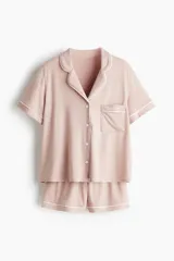 Pijama corto de dos piezas color rosado, en tejido de punto suave con ribetes blancos en contraste. Incluye camisa de manga corta con cuello abierto, botones al frente y bolsillo superior, y short con cintura elástica.