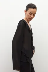 Blusa negra de manga larga, confeccionada en tela liviana y semitransparente, con escote en V adornado con volados y un botón frontal.