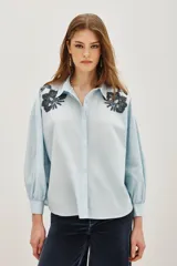 Camisa blanca de algodón con cuello clásico, mangas largas abullonadas con puños anchos y cierre frontal de botones. Presenta aplicaciones bordadas en dorado con detalles de animal print en los hombros.