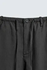 Pantalón negro de corte relajado y pierna ancha, confeccionado en mezcla de algodón y lino. Presenta cintura elástica ajustable con cordón y cierre frontal con cremallera y botón.