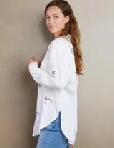 Camisa blanca con bordados coloridos y corte relajado, con botones frontales y dos bolsillos en el pecho.