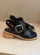 Sandalia negra de cuero vacuno con tiras cruzadas y hebilla metálica plateada grande. Posee un taco bajo de madera y suela de cuero.