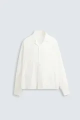 Camisa blanca de corte relajado confeccionada en tejido de algodón y lino. Tiene cuello bowling, manga larga con puños abotonados, jaretas en el delantero, bolsillos de parche en pecho y cadera, bajo con aberturas laterales y cierre frontal con botones.