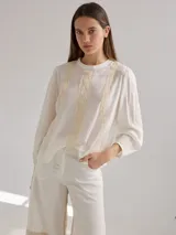 Camisa blanca de lino y viscosa, de corte holgado, con cuello redondo y mangas largas abullonadas. Presenta detalles de puntilla de algodón en el frente.