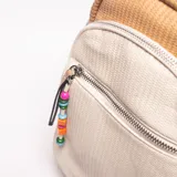 Mochila pequeña de estilo veraniego, combinando texturas tejidas en color beige claro y marrón. Presenta un bolsillo frontal con cierre y un colgante decorativo de cuentas de colores.