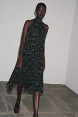 Vestido midi negro con estampado de lunares blancos, cuello halter con pañuelo integrado, sin mangas y bajo asimétrico. Cierre con abertura y botón en la nuca.