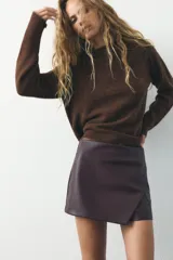 Falda pantalón corta marrón de tiro medio con efecto piel, falsos bolsillos de vivo en la espalda y cierre lateral con cremallera oculta.