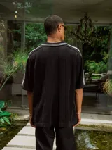 Chomba de tejido de punto cerrado color blanco, con cuello polo y mangas cortas. Presenta un bordado sutil en el frente y tres franjas negras en los hombros.