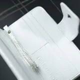 Billetera blanca con textura croco, cierre con broche y correa de mano.