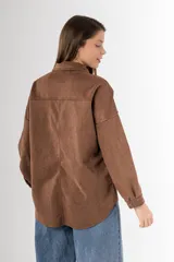 Camisa marrón de gamuza sintética, corte amplio y largo irregular. Tiene cuello clásico, cierre frontal con botones a presión, dos bolsillos con solapa abotonada en el pecho y mangas largas con puños abotonados.