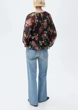 Blusa de mujer con estampado floral en tonos rojos, rosas y verdes sobre fondo negro. Tiene cuello redondo, mangas largas abullonadas, volantes en el bajo y cierre de lazo en el cuello.
