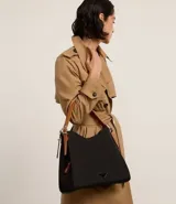 Cartera hobo grande color negro, confeccionada en poliéster y poliuretano. Cuenta con cierre de botón a presión, separadores con cremallera, bolsillo externo, asa mediana ajustable contrastante y detalle triangular de metal.