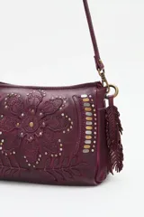 Cartera de cuero negro con diseño de flor mandala en relieve y apliques de tachas doradas y plateadas. Presenta flecos colgantes en uno de los laterales y correa ajustable para llevar al hombro o cruzada.
