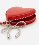 Billetera mediana roja con forma de corazón, cierre de cremallera, correa de mano y moño de perlas artificiales. Marca Satinato, colección Primavera/Verano 2025.