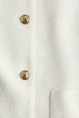 Chaqueta corta color crema en tejido texturizado con cuello clásico y botones metálicos decorativos. De corte ajustado con mangas raglán al codo y bolsillos de parche delanteros. Forrada.