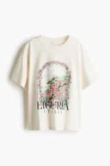 Remera de corte amplio color crudo, con cuello redondo y mangas cortas. Estampa frontal con dibujo de paisaje italiano y flores, y texto "Liguria Italia".