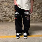 Pantalón de jean negro de corte super baggy con bordados de logos de marcas de automovilismo y texto en blanco y rojo.