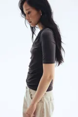 Remera negra de punto crepé con escote redondo y mangas cortas. Diseño ajustado con frunces en un hombro y en el lado opuesto de la cintura para crear un leve efecto drapeado.