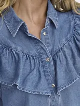 Camisa de denim celeste con cuello clásico, cierre frontal con botones dorados y mangas largas. Presenta volados en el frente y los hombros.
