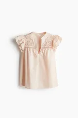 Blusa color rosa pálido de corte holgado en tejido liviano de algodón. Canesú con broderie adelante y liso atrás, ambos con frunces. Mangas cortas con volados.