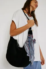 Bolso tipo hobo de gamuza negra, con forma holgada y cierre principal de cremallera. Se lleva al hombro con dos asas de correa delgadas sujetas con argollas metálicas plateadas.