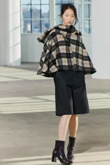 Capa de cuello alto confeccionada en tejido con lana a cuadros en tonos beige, negro y marrón. Tiene bajo asimétrico y cierre en la espalda con botón.