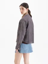 Chaqueta de cuero ovino color gris con terminación símil denim, de espíritu utilitario y moderno. Tiene bolsillos plaqué sobre el lado izquierdo y en cada lateral. El acceso es mediante broches metálicos. El cuello es de gamuza color tostado a contratono.