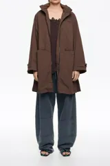 Gabardina midi de corte línea A en color bordó, confeccionada en mezcla de algodón técnico. Presenta cierre frontal con cremallera metálica, cuello subido con capucha integrada y mangas largas ajustables con botones de presión en los puños. Incluye bolsillos laterales y aberturas con botones en los costados.