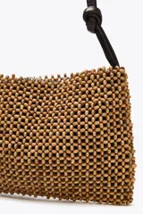 Bolso de hombro con cuerpo de abalorios color madera y asa de cuero sintético marrón con detalle de nudos. Tiene un bolsillo interior y cierre con cremallera metálica.
