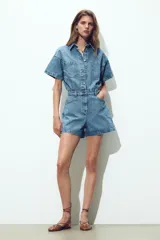 Mono corto de denim azul, con cuello solapa, manga corta, bolsillos delanteros y traseros, cintura elástica en la espalda y cierre frontal con cremallera y botones metálicos.
