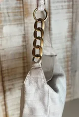 Cartera tipo hobo color blanco hueso con textura símil piel de cocodrilo, asa corta de cuero con eslabones de cadena dorada y cierre superior con broche imantado.