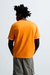 Camiseta naranja de algodón con cuello redondo y manga corta con vuelta. Estampado frontal con la firma del artista Otis Kwame Kye Quaicoe.