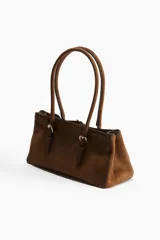 Cartera marrón de gamuza sintética con doble asa corta, cierre magnético y correa decorativa con hebilla metálica.