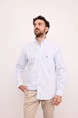 Camisa celeste con rayas verticales blancas, de corte clásico, con cuello abotonado, manga larga y bolsillo en el pecho.