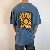 Remera azul con efecto gastado, corte oversize y estampa trasera con un sol amarillo dentro de un cuadrado negro y texto "This is living" y "Sunsets sessions".