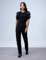 Pantalón negro de tiro alto y corte recto, confeccionado en tejido elastizado. Presenta un diseño frontal con botones decorativos aplicados en los laterales de la cintura y costuras verticales que estilizan la silueta.
